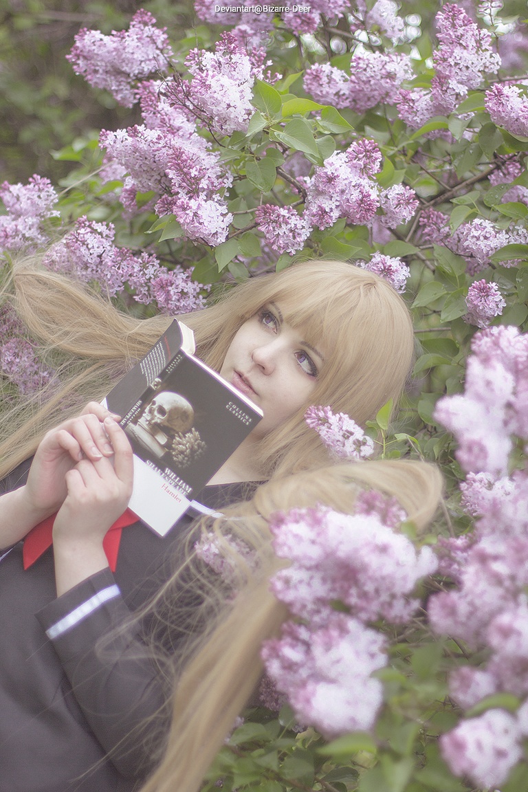 A person with long blonde hair dressed in a black outfit, holding a book titled 'Self-Portrait' while surrounded by purple lilac flowers.