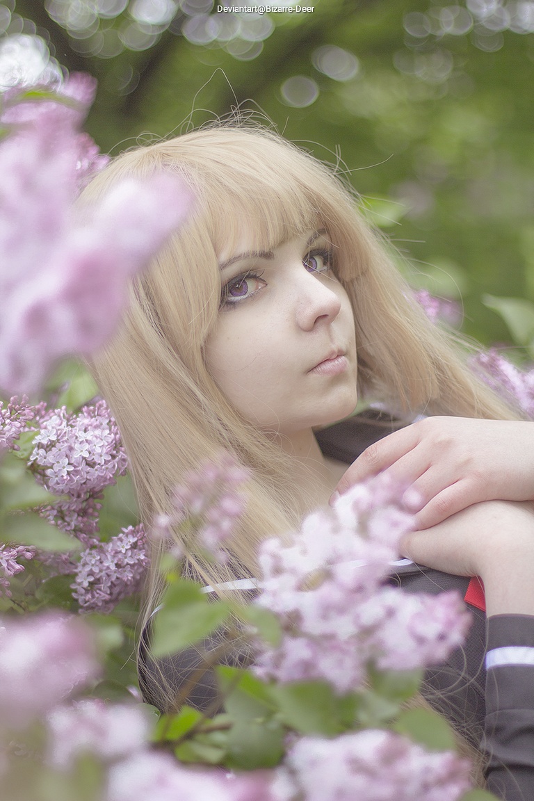 A young person with long, blonde hair poses amongst blooming lilac flowers, wearing a dark outfit. The background is softly blurred, emphasizing the subject's expression.