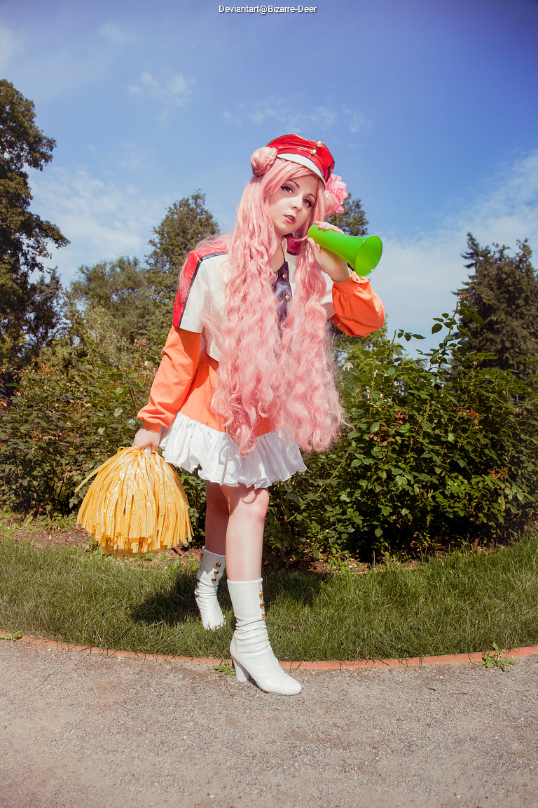 A person with long pink hair dressed in a colorful cheerleader-style outfit, holding a green megaphone and a yellow pom-pom, stands in a park with trees and blue sky in the background.