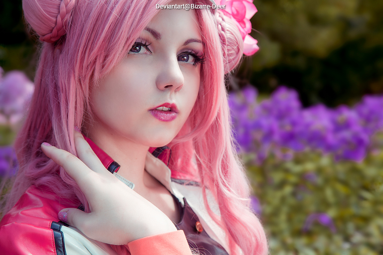 A close-up portrait of a person with long pink hair styled in two buns, wearing a colorful outfit. The background features purple flowers, creating a vibrant and visually appealing scene.