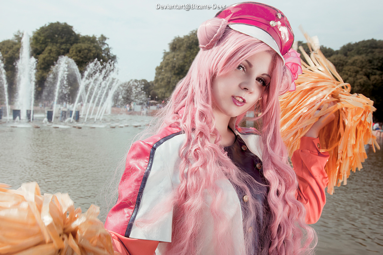 A young woman with long pink hair wearing a cap and holding pom-poms stands in front of a fountain in a park.