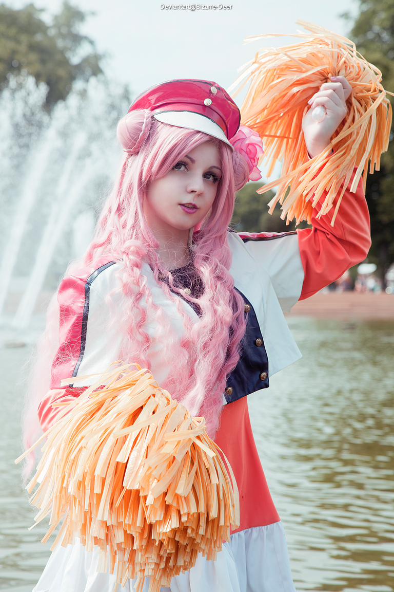 A person with long pink hair wearing a red and white cheerleading outfit, holding cheerleader pom-poms, posing in front of a fountain.