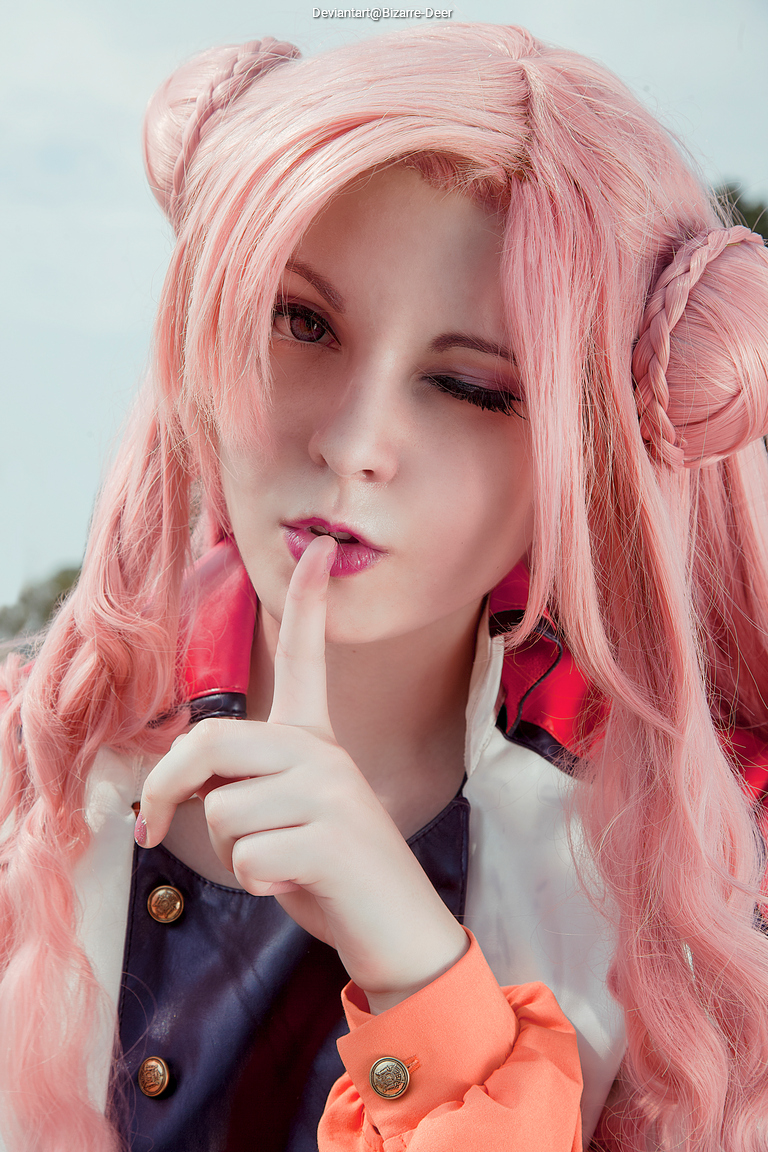 Close-up portrait of a person with long pink hair styled in buns, making a 'shh' gesture with their finger to their lips.