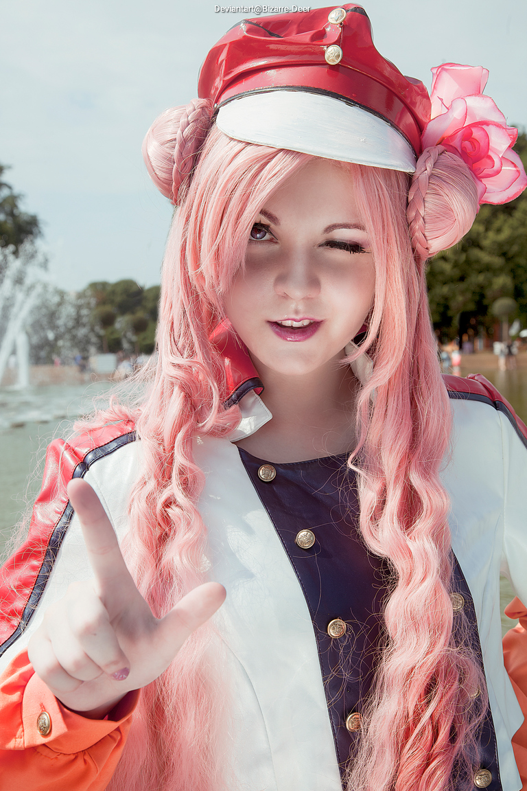 A person in a colorful costume with pink hair, wearing a red cap and winking while pointing with one hand.