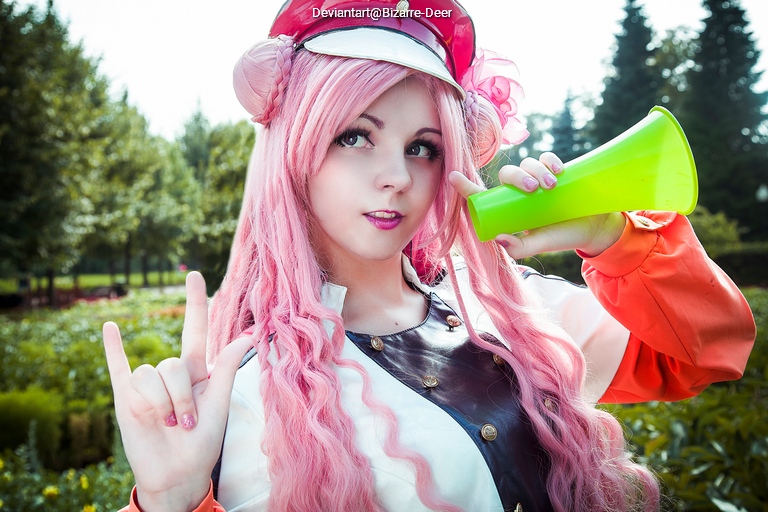 A person with long pink hair, wearing a cap and a white jacket, holds a green megaphone and makes a hand gesture while standing in a garden.