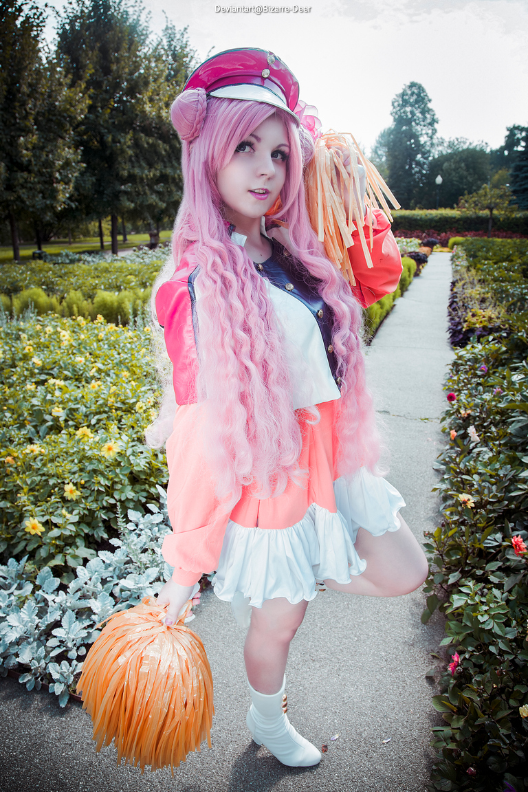 A person with long pink hair wearing a colorful outfit and a hat, holding orange pom-poms in a garden setting.