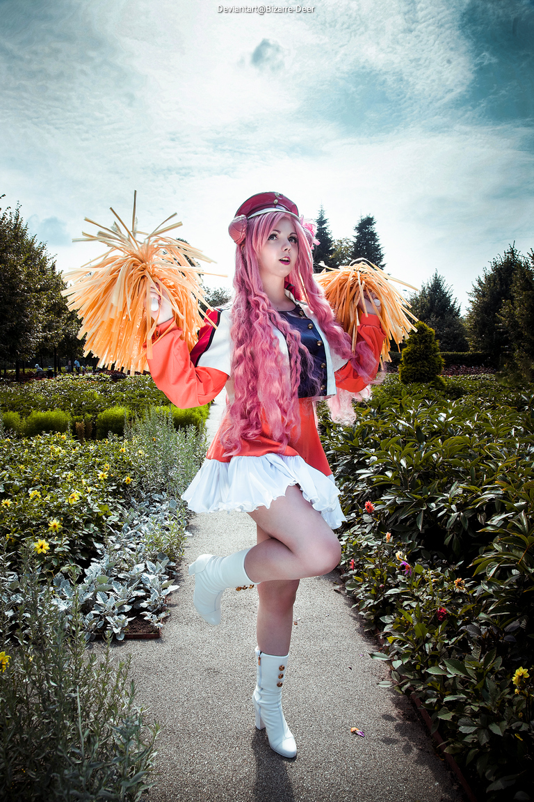 A person with long pink hair wearing a cheerleader outfit, holding pom-poms, posing in a garden setting with flowers and greenery.