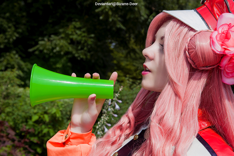 A person with long pink hair is holding a bright green megaphone to their mouth. They are in an outdoor setting with greenery in the background.