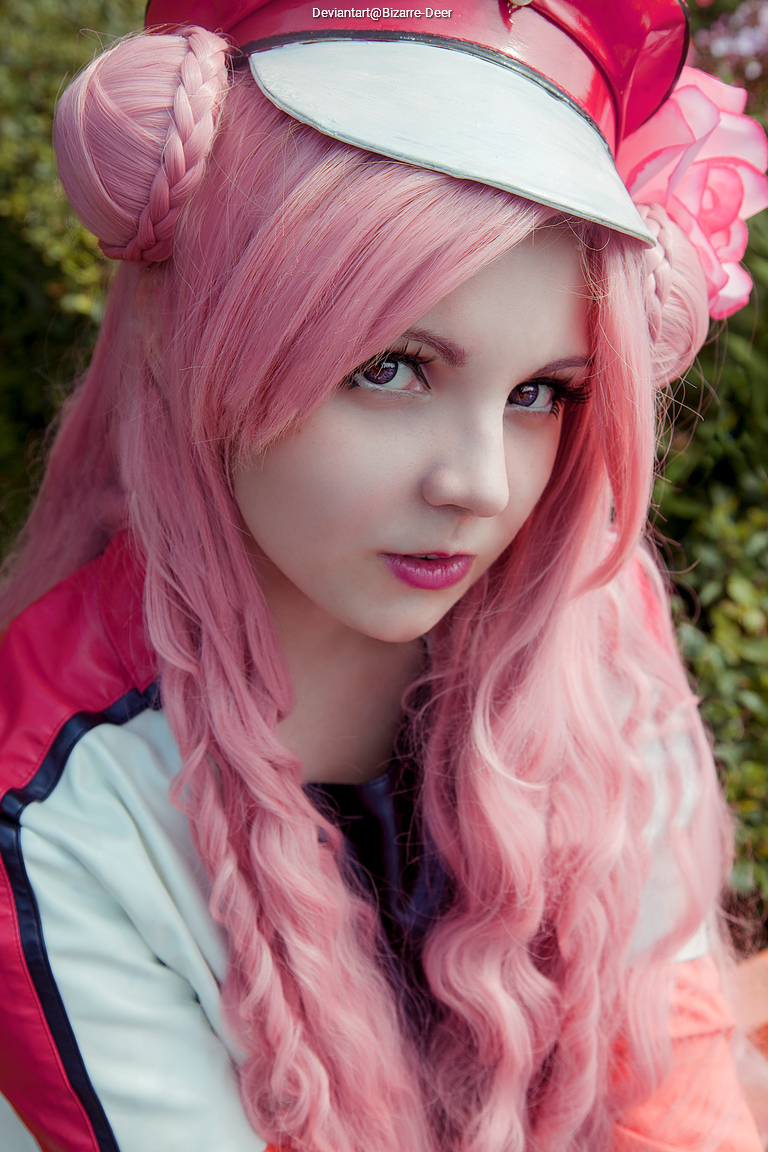 Close-up portrait of a young woman with long pink hair styled in braids, wearing a red cap and a colorful jacket, surrounded by greenery.