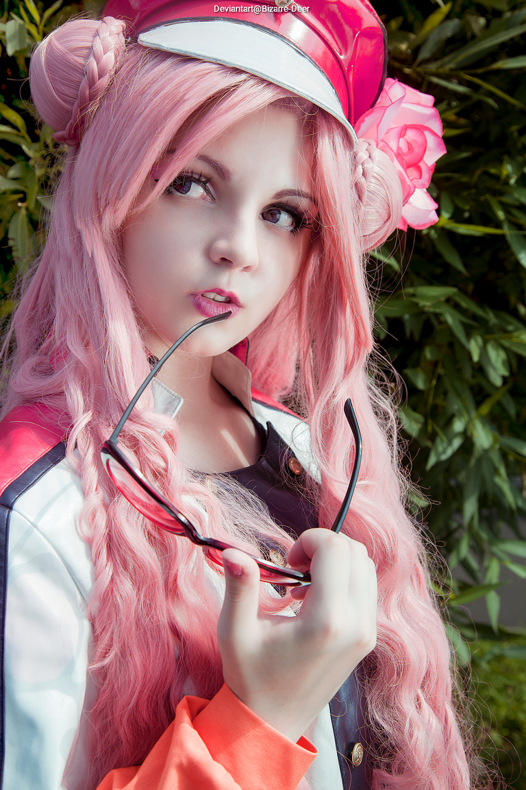 A person with long pink hair styled in buns, wearing a red cap and a colorful jacket, holding sunglasses in their mouth while standing in front of greenery.