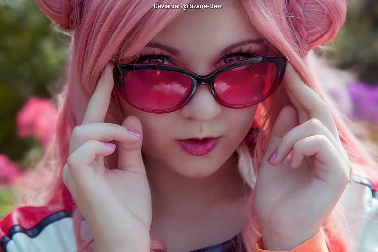 A close-up portrait of a person with pink hair and pink sunglasses, striking a confident pose against a blurred floral background.