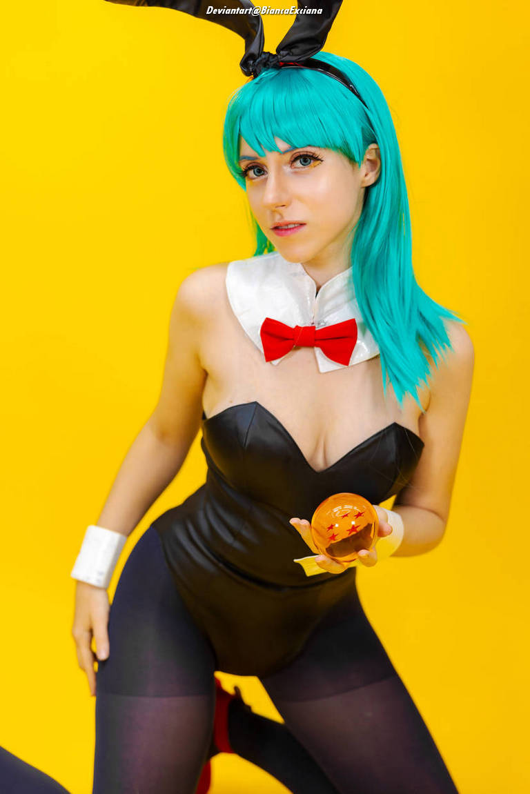 A person with teal hair wearing a black bunny outfit and a bow tie, holding a Dragon Ball, set against a bright yellow background.