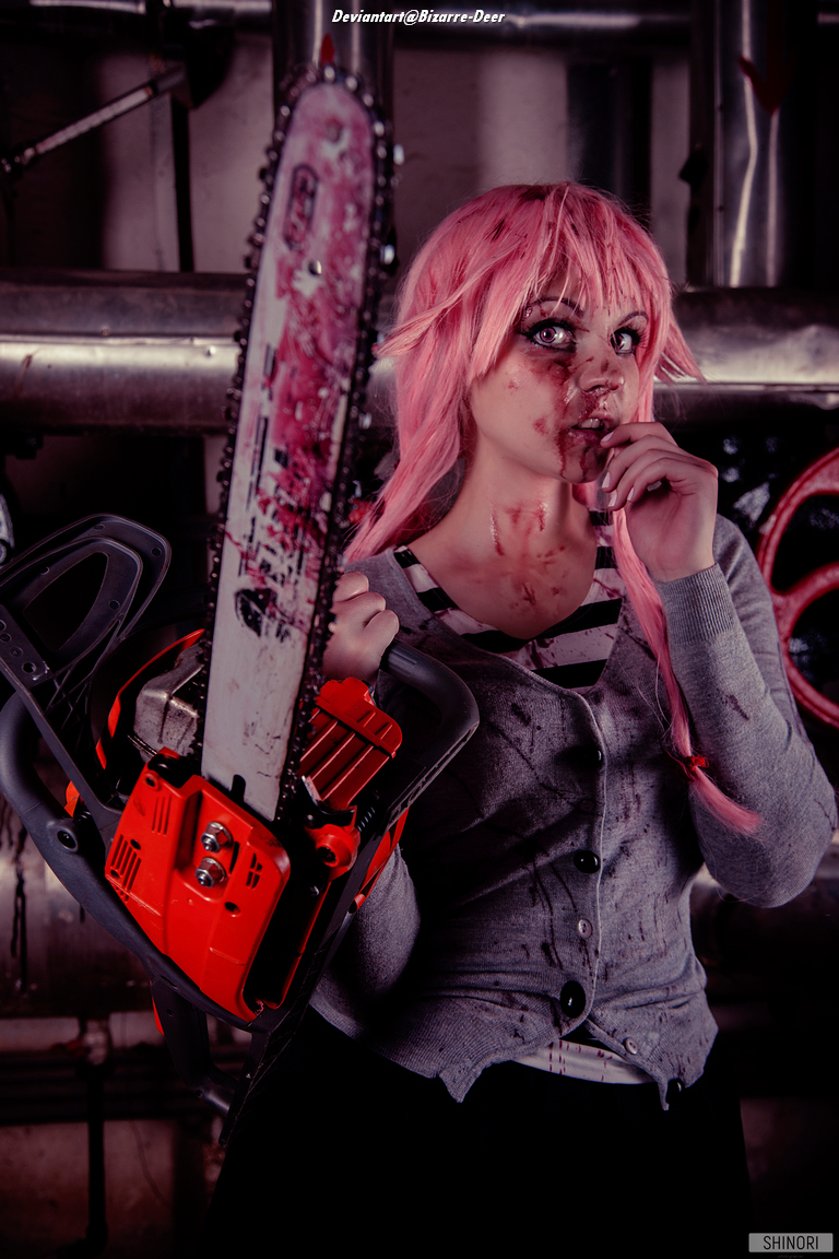A person with pink hair and a gray cardigan holds a chainsaw in a dimly lit industrial setting, with fake blood on their face and clothes.