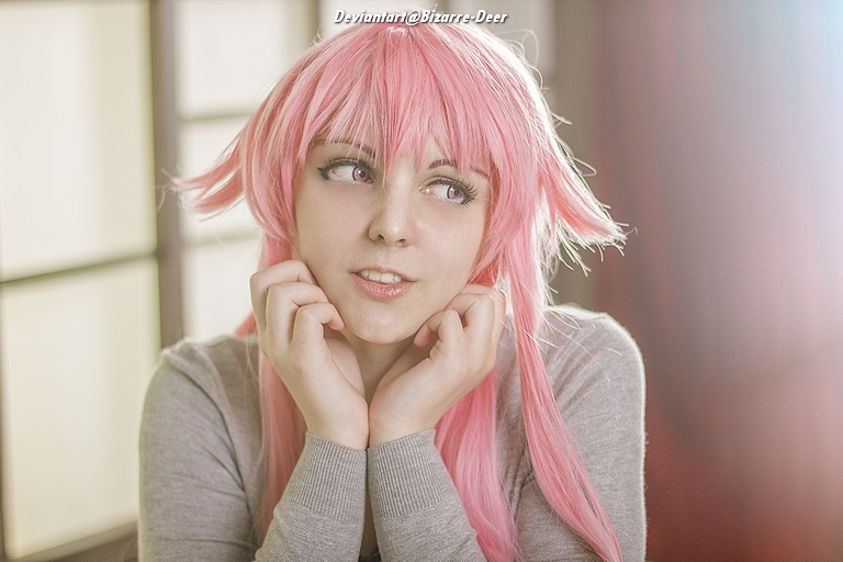 A person with pink hair, wearing a gray sweater, posing thoughtfully with their hands under their chin in a softly lit interior.