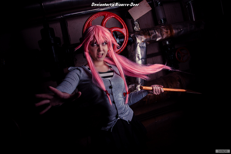 A person with long pink hair wearing a gray cardigan and holding a paintbrush, posing dramatically in a dimly lit industrial setting.