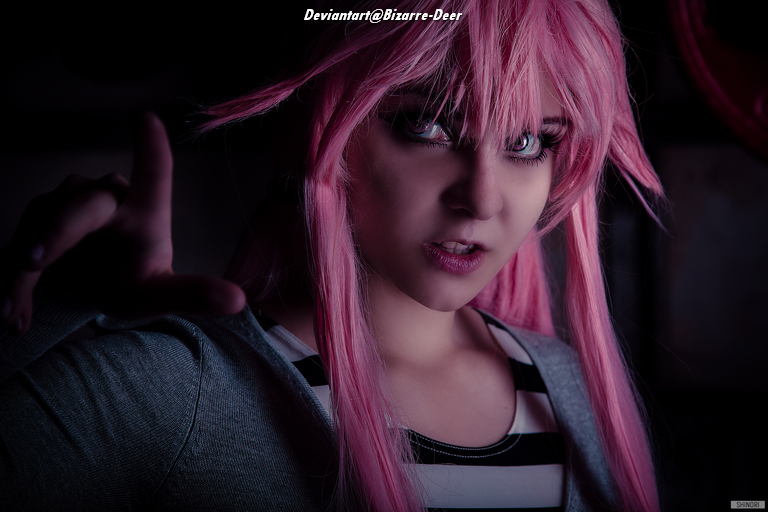 A person with pink hair and striking makeup, posing dramatically with one hand raised, dressed in a striped top against a dark background.