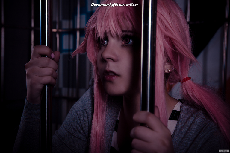 A young person with pink hair peering through metal bars, wearing a gray sweater and a striped shirt, in a dimly lit environment.
