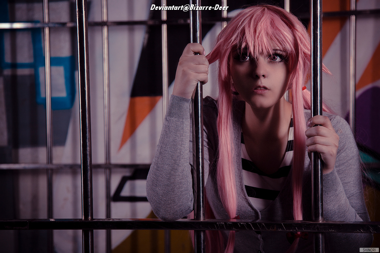 A person with pink hair is sitting behind bars, looking intently at the camera with a worried expression. They are wearing a striped shirt and a gray cardigan, with a colorful background.