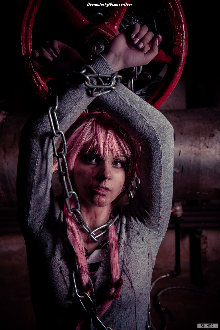 A person with long pink hair is chained and poses dramatically in a dimly lit industrial environment, with a large red valve behind them.