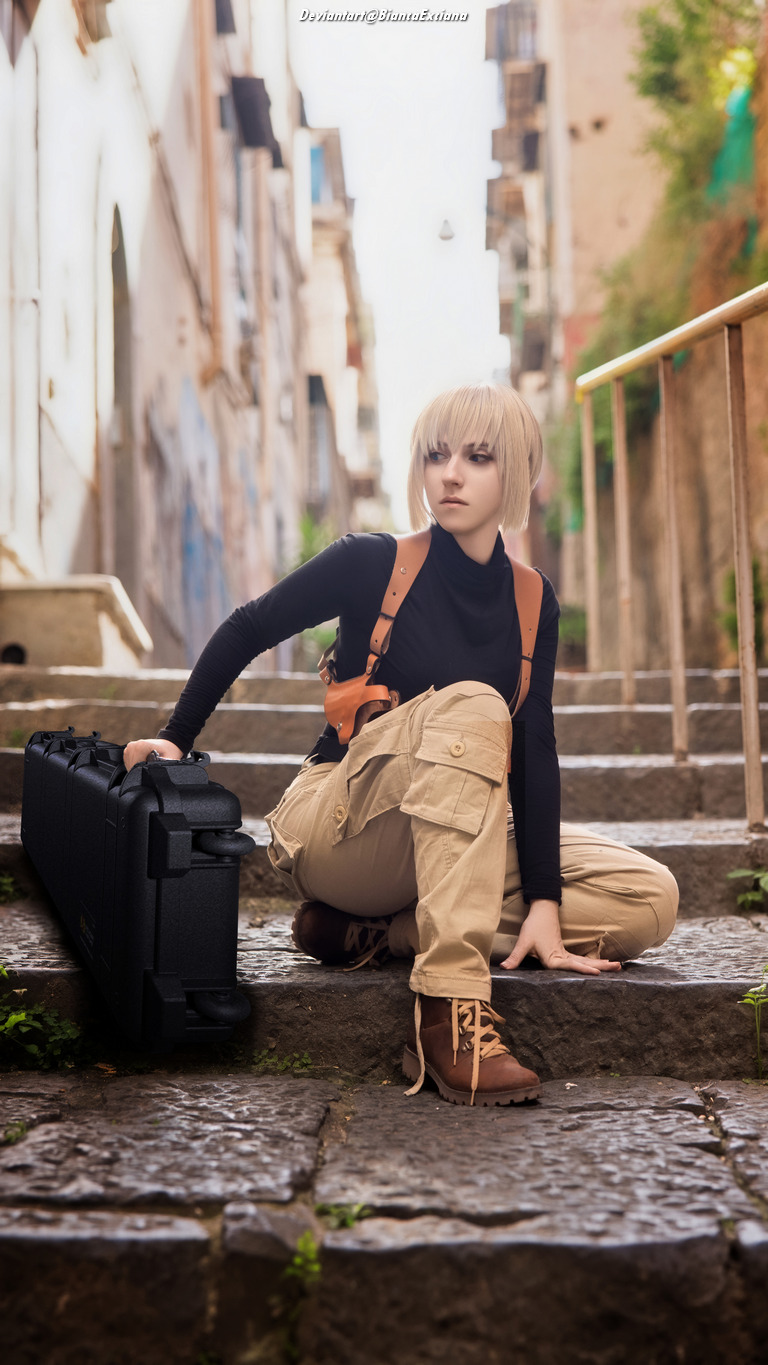 A person with short blonde hair dressed in a black long-sleeve shirt and beige cargo pants, sitting on stone steps with a black case beside them. They appear focused and stylish in an outdoor setting with buildings in the background.