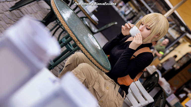 Person with short blonde hair sitting at a table, holding a cup while enjoying a drink. The setting appears to be a café with a casual atmosphere.