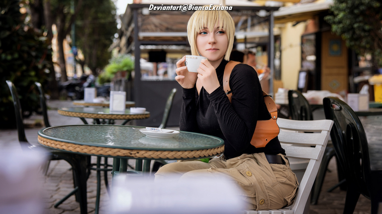 Person sitting at an outdoor cafe table, holding a cup, wearing a black long-sleeve shirt, and light-colored pants.