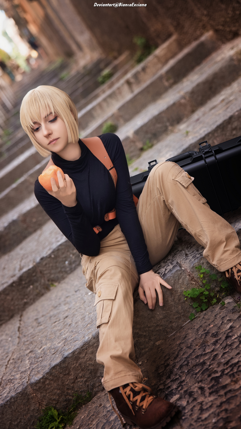 A person with short blonde hair dressed in a black long-sleeve shirt and beige cargo pants sits on stone steps, holding a piece of fruit. They have a pensive expression, with a black bag beside them.