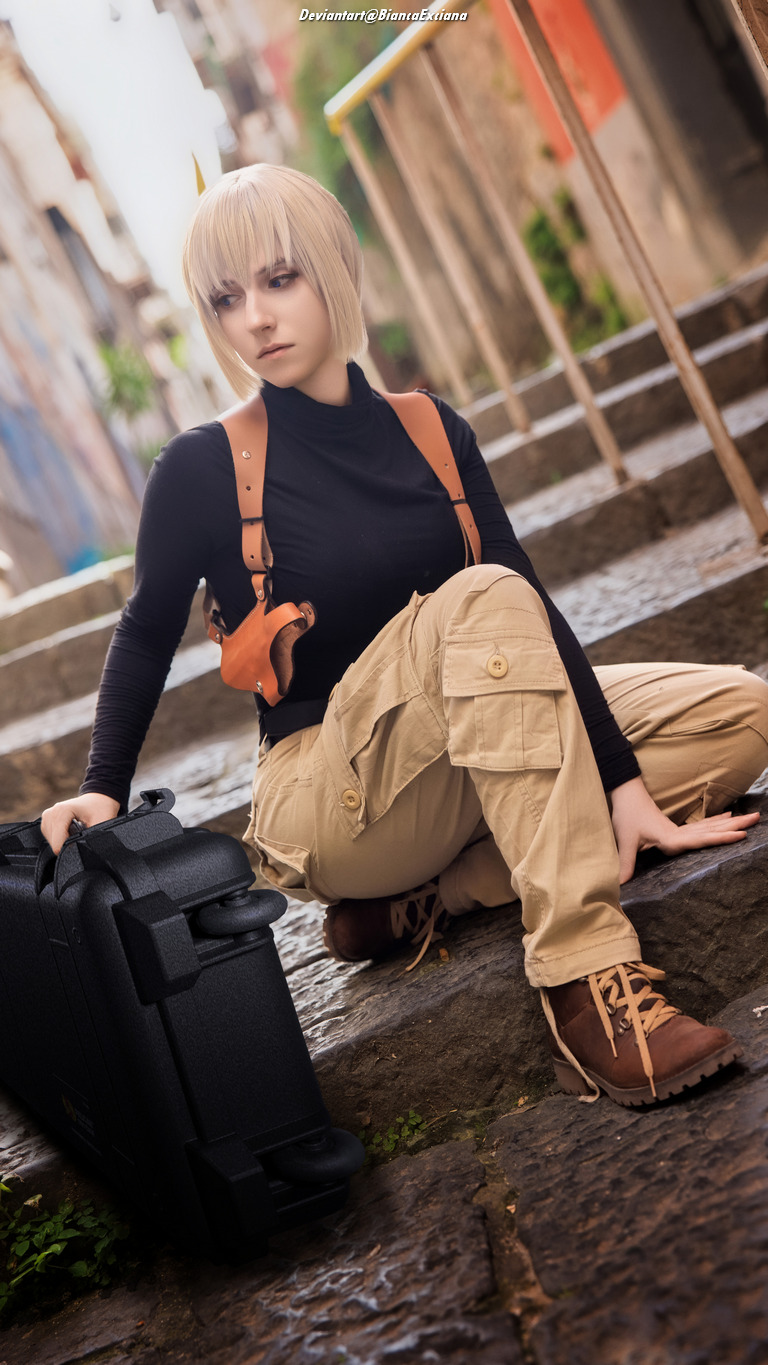 A person in a black top and beige cargo pants squats on a stone path, next to a black carrying case, with a backdrop of colorful buildings.