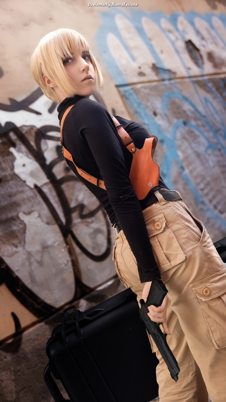 A person with short blonde hair dressed in a black long-sleeve top and tan cargo pants, holding a gun and standing next to a black case, with graffiti in the background.