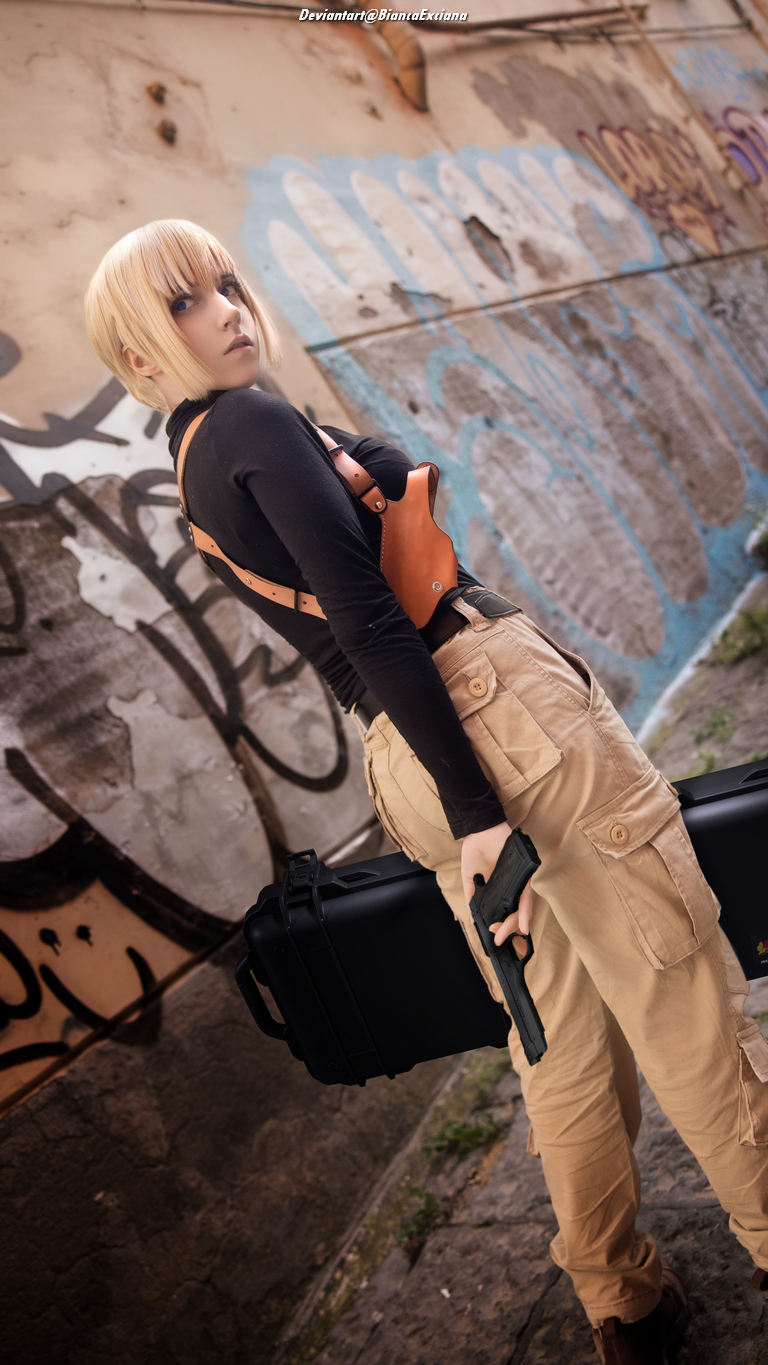 A character in urban cosplay fashion holding a gun and a black case, standing against a graffiti-covered wall.