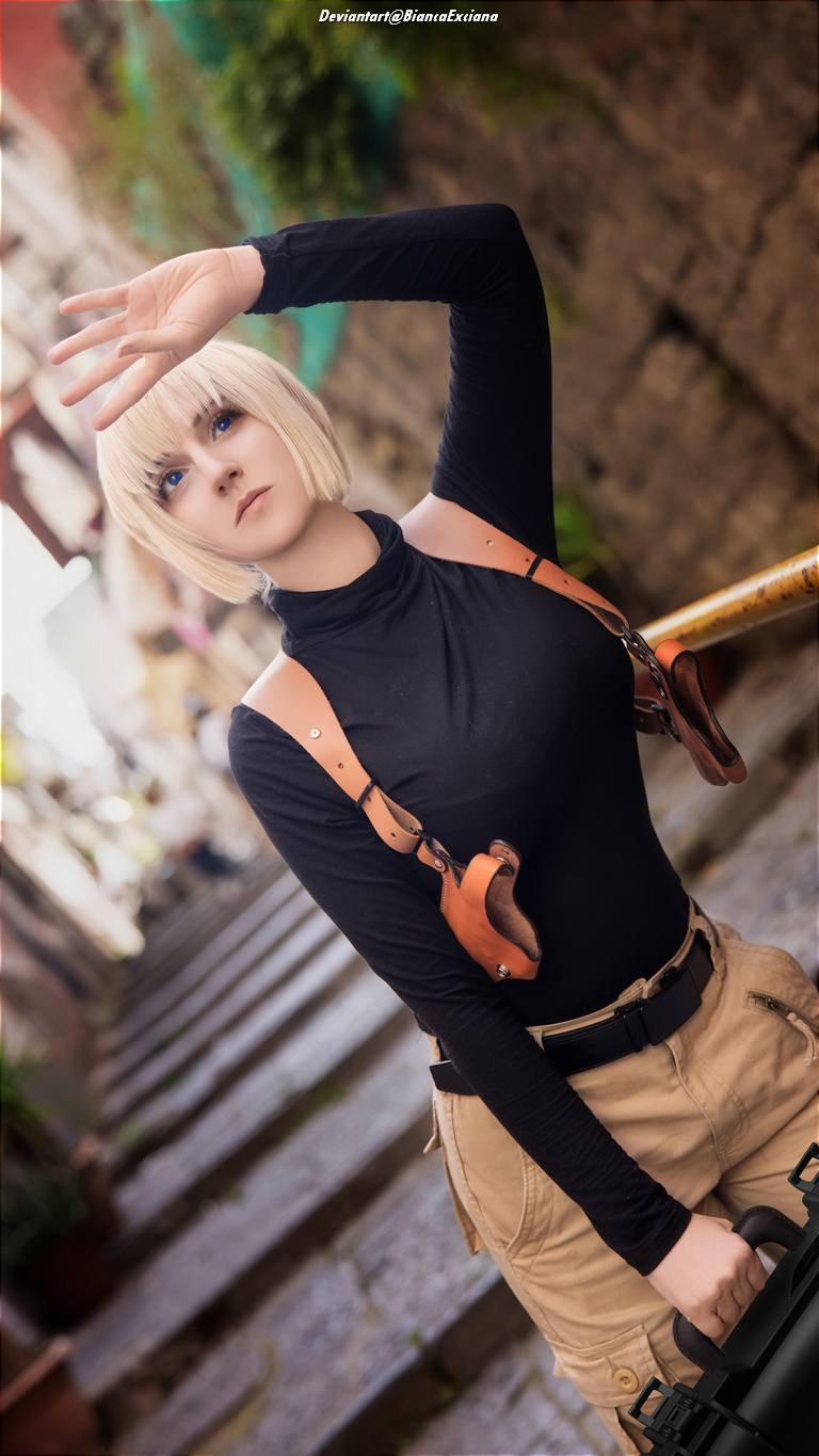 A person in a black long-sleeve top and beige shorts poses with one hand raised in a street setting, wearing a holster. They have short blonde hair and striking blue eyes.