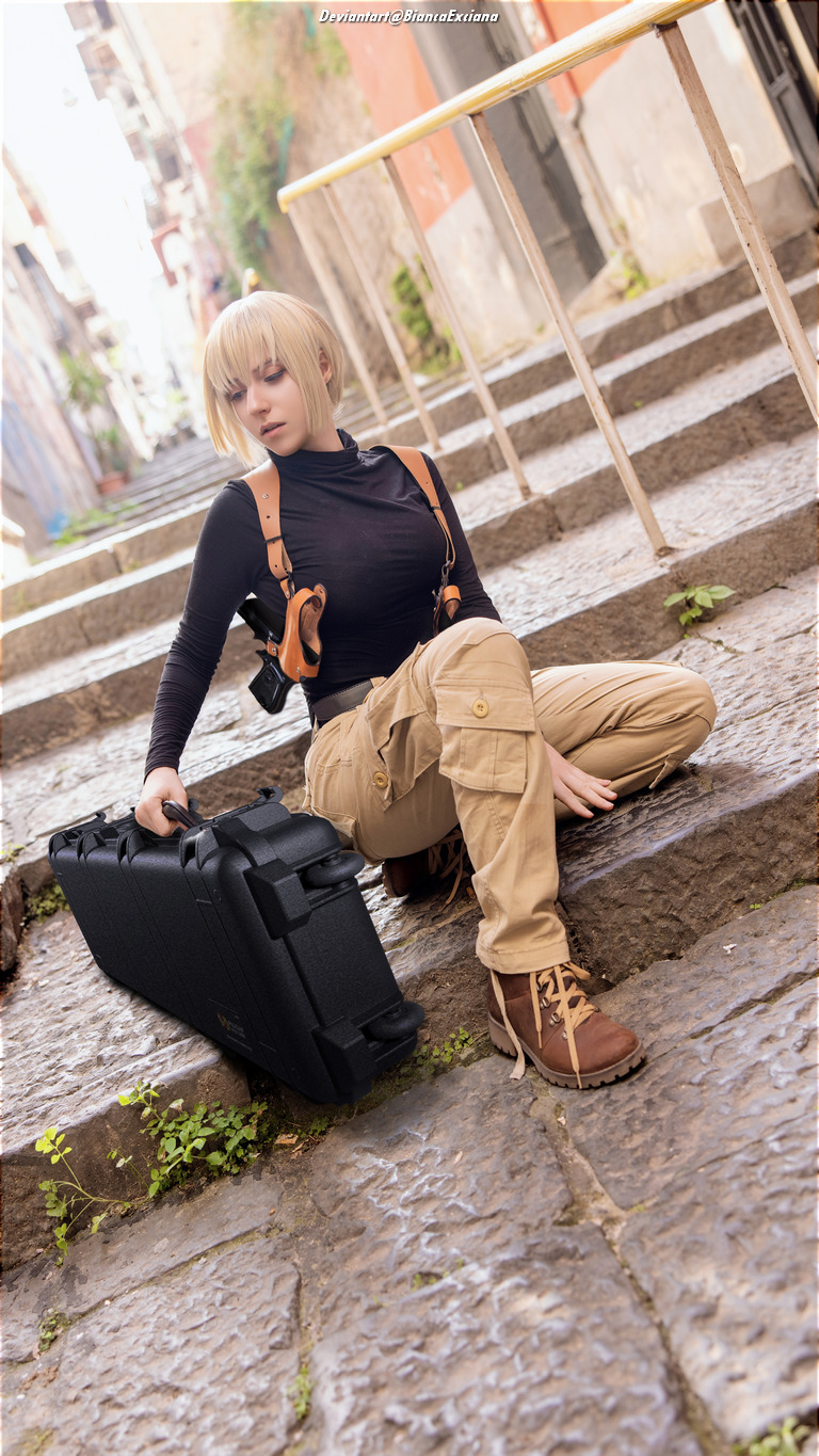 A person with short blonde hair, wearing a black top and tan cargo pants, sits on stone steps holding a black case, in a narrow alleyway.