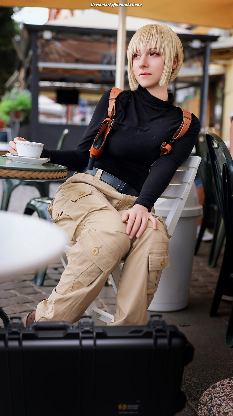 A person with short blonde hair sitting at an outdoor cafe, wearing a black top and beige cargo pants, with a cup of coffee on the table.