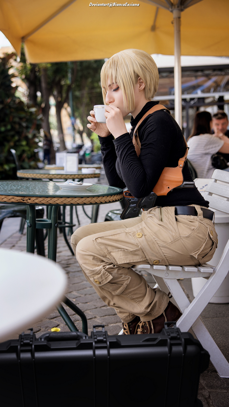 A person with short blonde hair is sitting at a table in a cafe, holding a small cup. They are wearing a black top and beige cargo pants, with a brown belt. A black case is positioned next to their chair.
