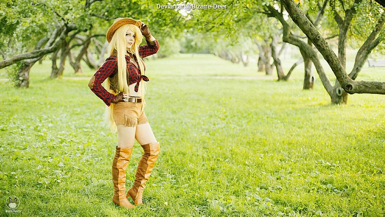 A person dressed in a western-style outfit with a cowboy hat, standing in a grassy field surrounded by trees.