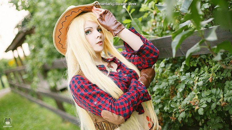 A person in a cowgirl outfit with long blonde hair and a plaid shirt poses outdoors among green foliage.