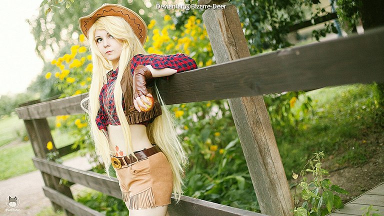 A cosplayer dressed as a character with long blonde hair, wearing a cowboy hat, a plaid shirt, and tan shorts, posing by a wooden fence in a garden with sunflowers in the background.