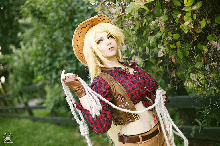 A person dressed in a cowboy costume with a plaid shirt and a cowboy hat, posing outdoors while holding a rope.