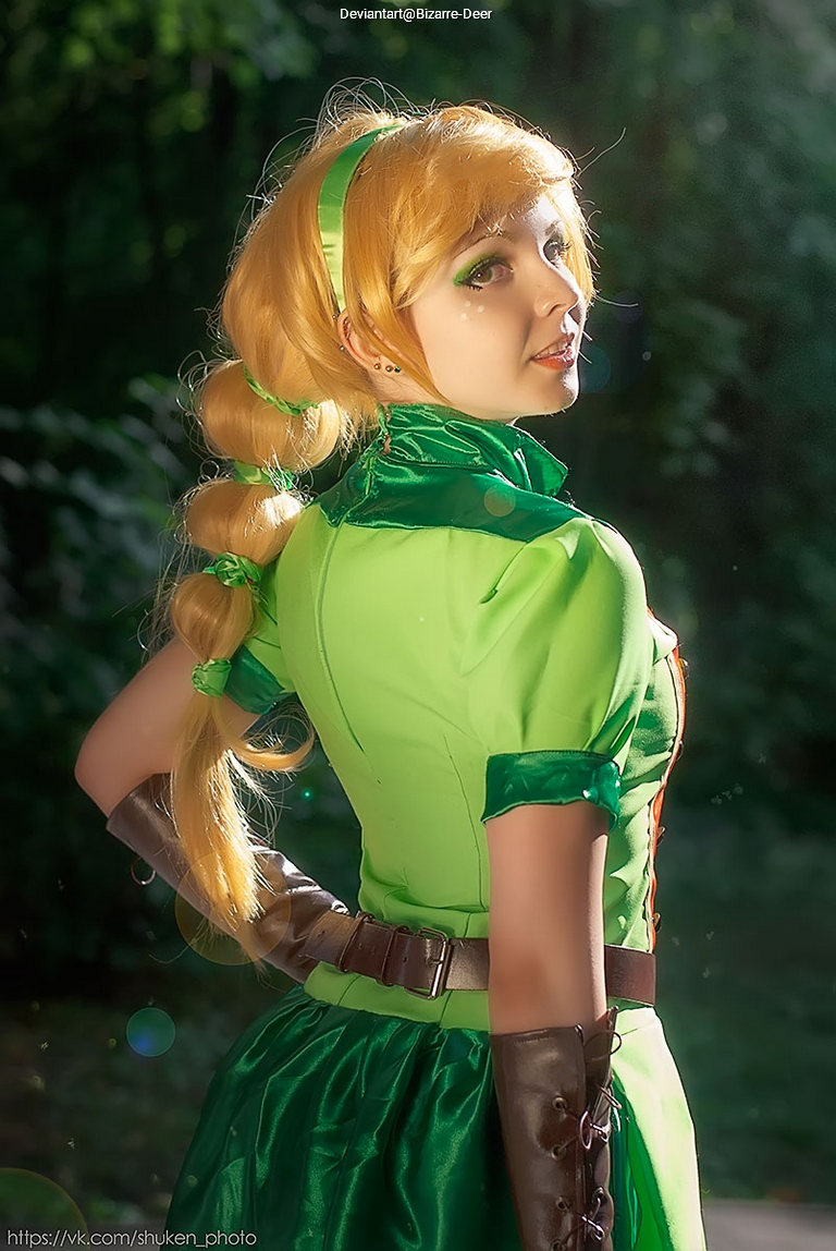 A person in a green costume with blonde hair styled in braids, standing outdoors and looking back over their shoulder, with soft lighting highlighting their features.