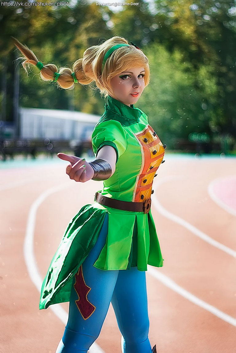 A person in a green and yellow cosplay outfit with braided hair stands on a running track, extending one hand forward.