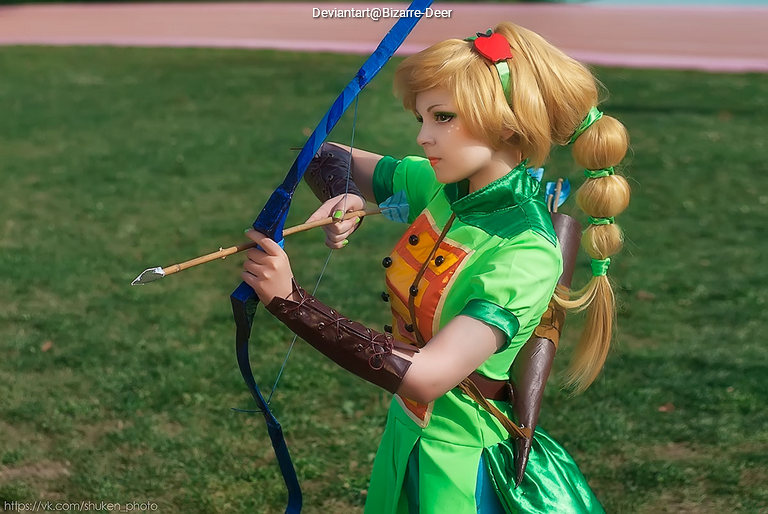 A person dressed in a green costume, holding a bow and arrow, posed in a grassy area.
