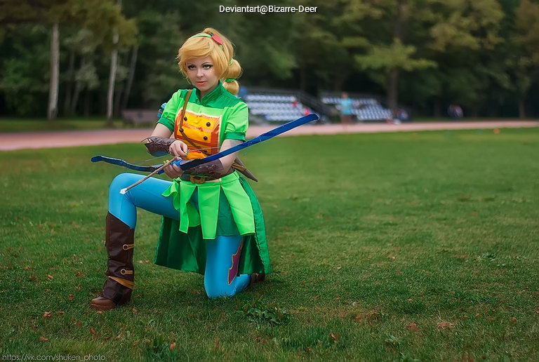 A person dressed as a fantasy character in a green outfit with a bow, kneeling on grass in a park.