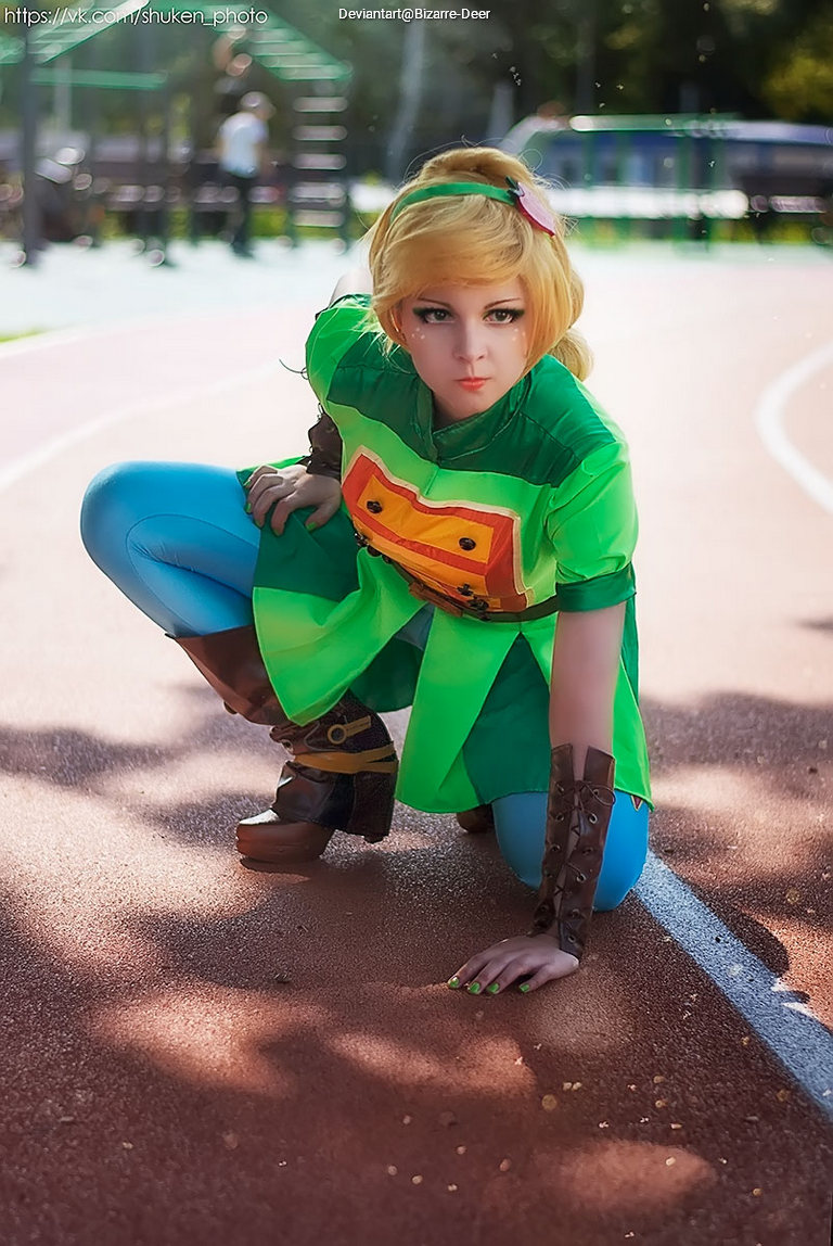 A person dressed in a green fantasy costume with a blonde wig, crouching on a track surface, appearing focused and ready to sprint.