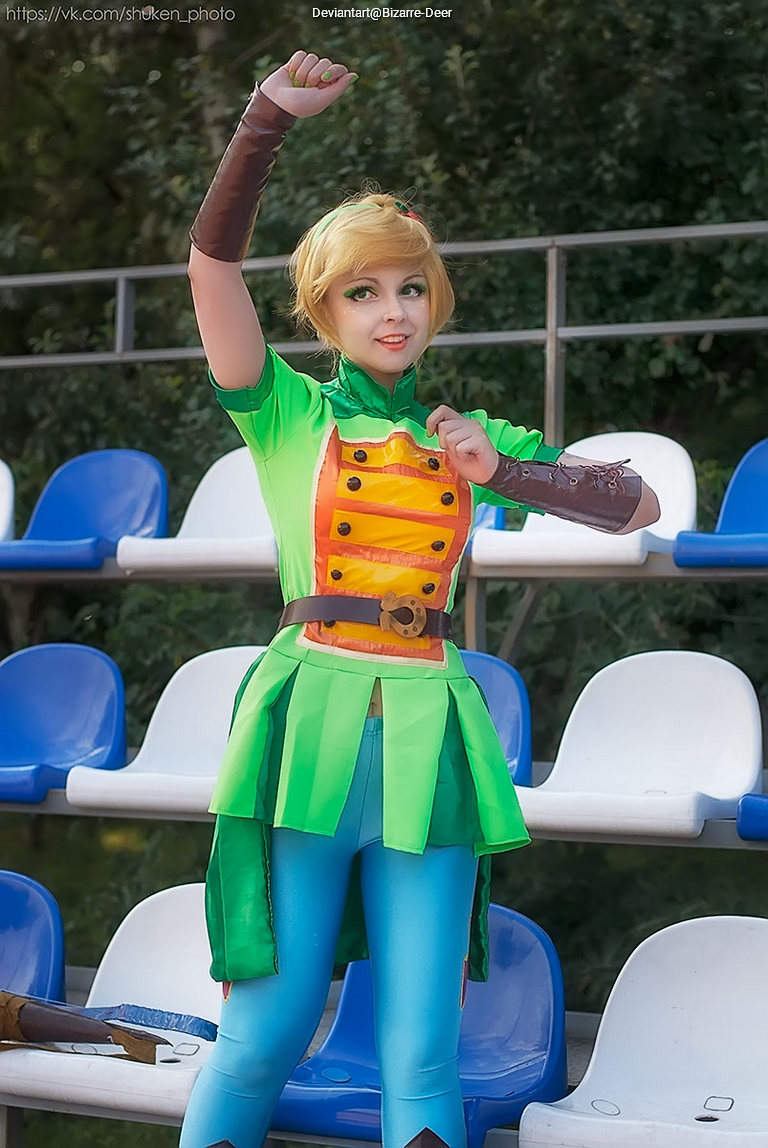 Person in a colorful green costume with a yellow design, posing with one arm raised in a lively manner, set against a backdrop of empty bleachers.