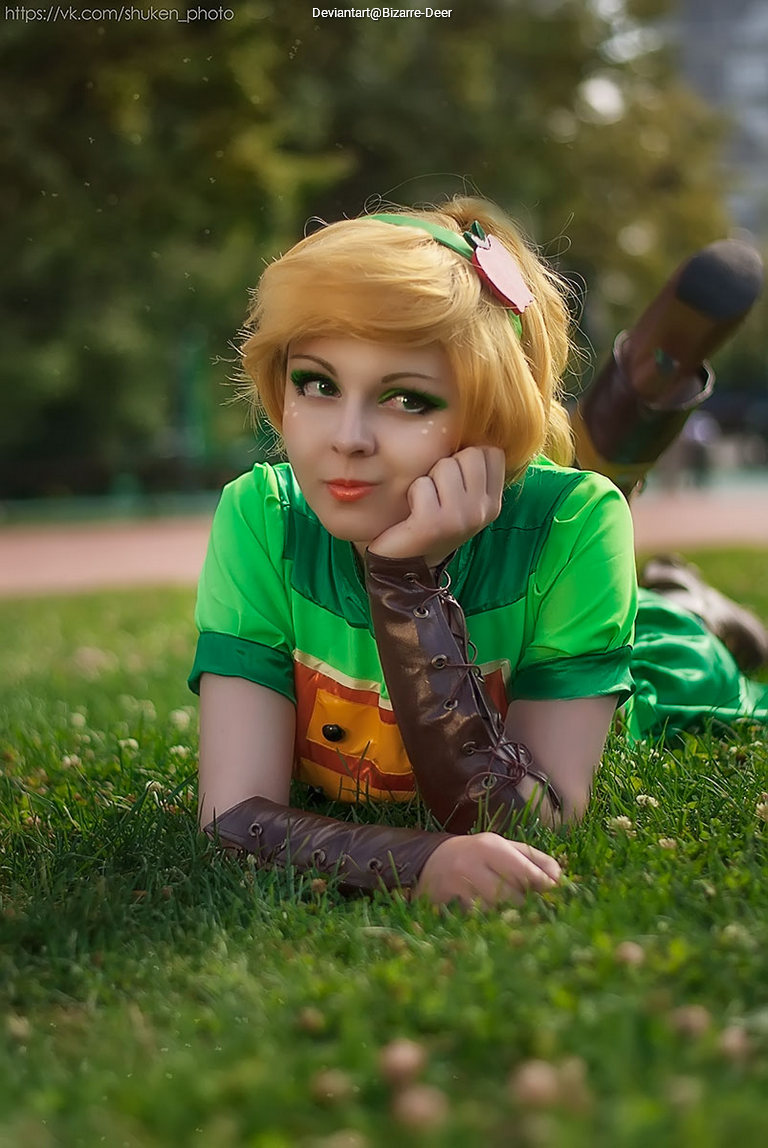 A person dressed in a vibrant green costume lies on grass, resting their head on their hand and looking playfully towards the camera.