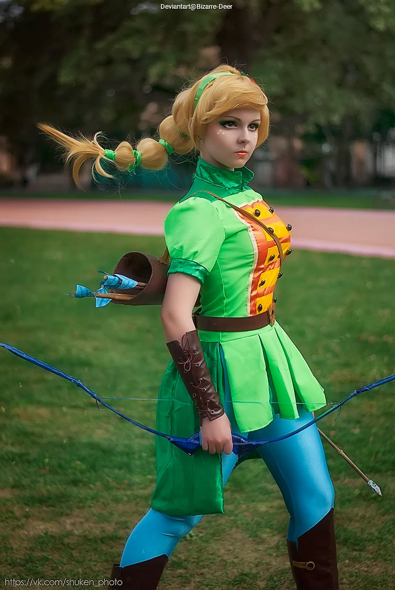A cosplayer dressed as an archer, wearing a green costume with blue leggings, holding a bow, and standing in a park setting.