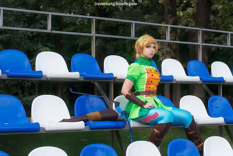 A person dressed as a fantasy character sits alone on blue and white bleacher seats in an outdoor setting, wearing a green outfit with blue pants and brown accessories.