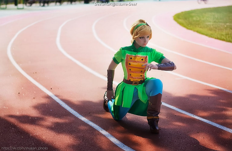 Person in green and brown costume crouched on a running track, posing dramatically.
