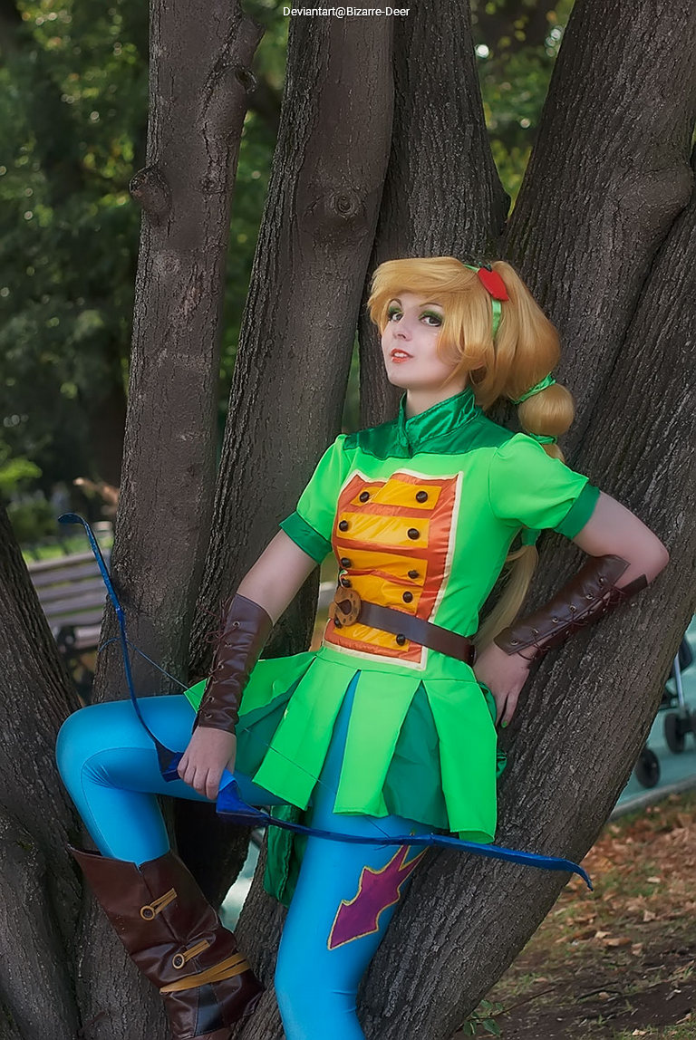Person in a green costume with a bow and arrow, posing against a tree.