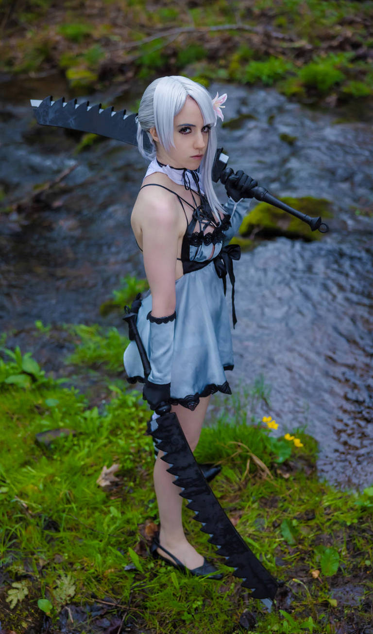 A person in a unique costume stands by a stream, holding a large, intricately designed sword. They have long silver hair and a flower accessory, and the scene is surrounded by lush green foliage.